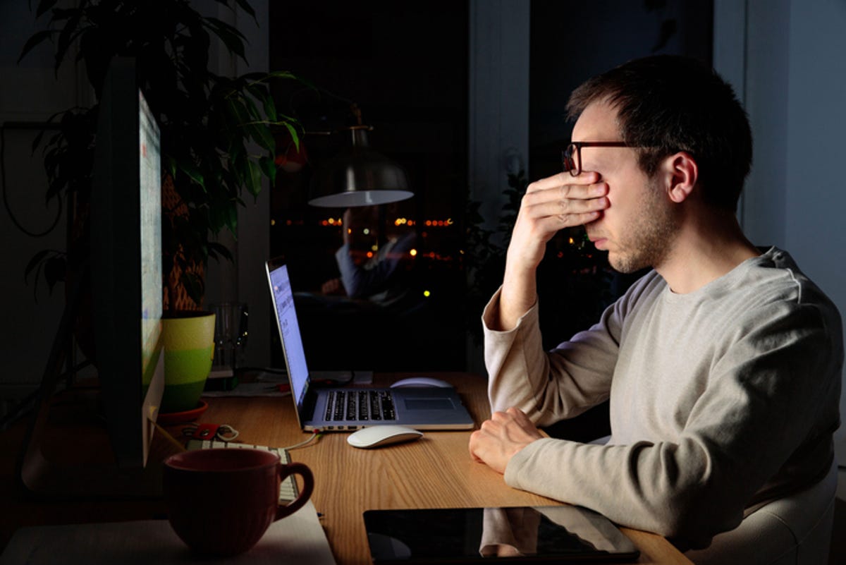 Homem sentado em frente a um computador no escuro, esfregando os olhos.
